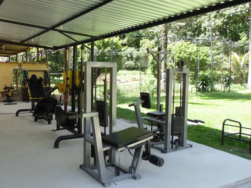 Aparelhos de musculação sob cobertura de telhas metálicas, ao lado de um gramado cercado por tela